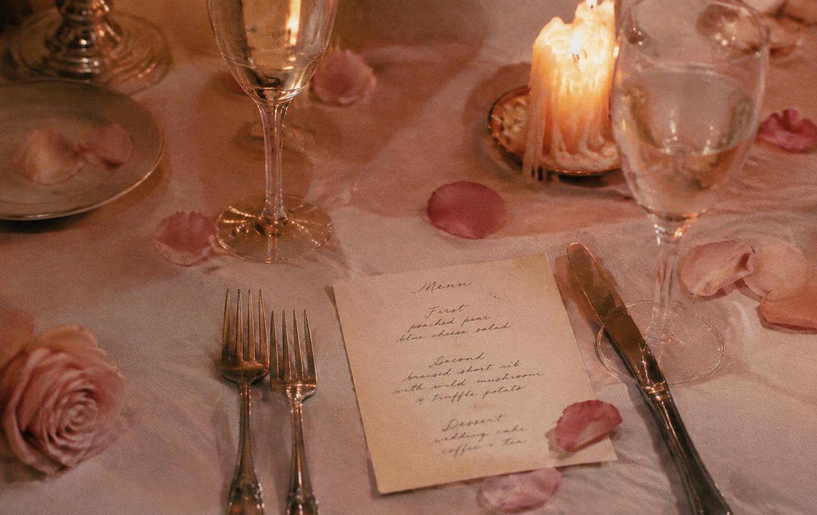 A handwritten menu, champagne glasses, rose petals and candle wax on linen